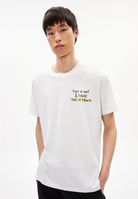 Witte katoenen t-shirt met zwart en gele tekst: "DIT IS GEEN T-SHIRT. DIT IS GEAFFIGEERD." Korte mouwen en ronde halslijn.