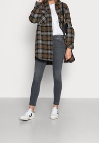 Abrigo de cuadros de gran tamaño en marrón, gris y naranja, combinado con jeans ajustados grises y zapatillas blancas. Presenta bolsillos y una tela texturizada.