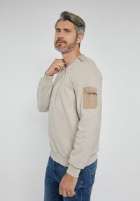 Beżowy sweatshirt z okrągłym dekoltem, teksturowanym materiałem i kieszenią na rękawie. Posiada kontrastowe akcenty na ramionach oraz ściągacze przy mankietach.