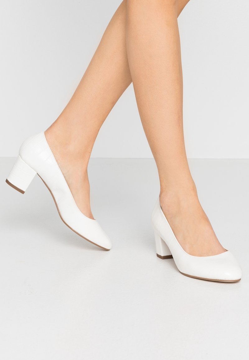 Jambes portant des chaussures blanches à talons moyens fermées, debout sur une surface gris clair avec un fond clair uni.