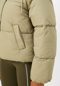 Vaalean khaaki puffer-takki, jossa on tikattu muotoilu, joustavat hihansuut ja sivutasku. Valmistettu sileästä, hieman teksturoidusta materiaalista.