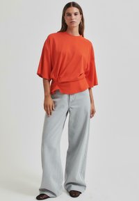 Oranžové oversized tričko s uzlem na přední straně, kombinované se světle šedými širokými džíny a černými síťovinovými botami.