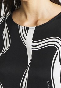 Svart topp med vita vågiga linjer och rund halsringning. Tyget verkar slätt med en lätt glans. Synlig sömnadsdetalj vid halsen.