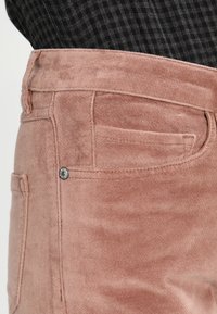 Detailní záběr na vybledlý růžový semišový kapsu a pas kalhot, které jsou nošeny s černým a šedým kostkovaným topem.