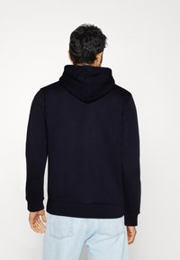 Felpa con cappuccio blu navy in un tessuto morbido, con tasca a marsupio e polsini a coste. Il modello indossa jeans blu chiaro.