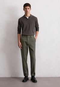 Chemise polo à manches longues gris foncé avec un col, assortie à un pantalon taillé vert olive et des chaussures noires, se tenant contre un fond neutre.