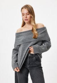 Dunkelgrauer, schulterfreier Pullover mit geripptem Halsausschnitt und Bündchen, kombiniert mit hochtaillierten schwarzen Jeans. Weicher Stoff, lockere Passform.