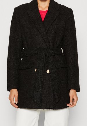 Femme portant un manteau noir texturé ceinturé sur un haut rouge et un pantalon blanc, debout les bras détendus le long du corps.