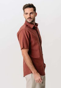 Mand iført en kortærmet rustfarvet skjorte med knapper og lysebeige bukser, der ser til sin venstre side mod en simpel hvid baggrund.