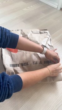 Barna papírcsomag egy világos padlón, két kéz rögzíti a csomagot, látható vonalkódokkal és QR-kóddal; texturált papírfelület.