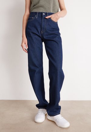 Person iført mørkeblå jeans med høj talje, med en ribbet ærmeløs top og hvide sneakers, stående op ad en ensfarvet væg.