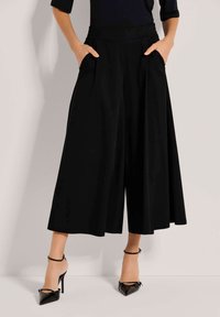 Zwarte brede broek met hoge taille, geplooide voorkant en zijzakken. Gecombineerd met glanzende zwarte hoge hakken.