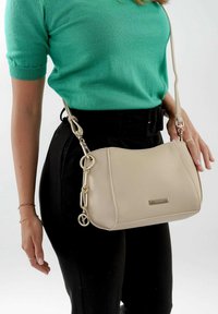 Beige Umhängetasche aus strukturiertem Leder mit goldenen Beschlägen und einer Kettenakzentuierung. Sie hat eine flache Form und einen verstellbaren Tragegurt, gehalten von einem Model.