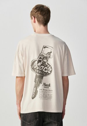 Mann trägt beige T-Shirt mit großem schwarzem Aufdruck eines Mack Trucks-Schlüssels und Bulldoggen-Logos auf der Rückseite.