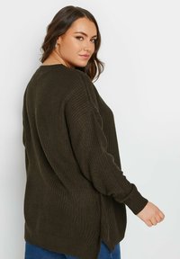 Mörkgrön, texturerad stickad tröja med långa ärmar, avslappnad passform, sidofickor och ribbade ärmslut, tillsammans med blå jeans.