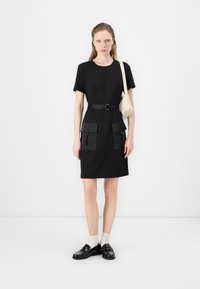 Black short-sleeve dress with a round neckline, belted at the waist, featuring two large front pockets. Worn with loafers and white socks.