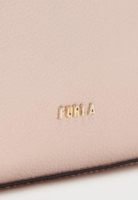 Κοντινό πλάνο επιφάνειας από υφή ροζ δερμάτινου με χρυσά μεταλλικά γράμματα της μάρκας "FURLA" και μαύρο περίγραμμα στο κάτω μέρος.
