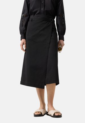 Kobieta w czarnej midi spódnicy typu wrap, czarnej bluzce z długim rękawem z koronkowymi wycięciami, beżowych sandałach, trzymająca okulary przeciwsłoneczne w prawej ręce.