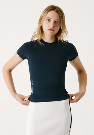 T-shirt ajusté à manches courtes navy en tissu lisse, arborant un logo imprimé sur le côté gauche. Associé à une jupe crayon blanche.