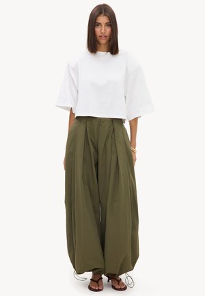 Femme portant un haut blanc ample à manches courtes et un pantalon plissé large vert olive avec cordons, debout et regardant vers l'avant sur fond blanc.