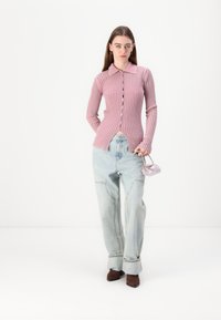 Cardigã de malha em rosa claro com gola e fecho de botão, combinado com jeans azuis claros largos e uma pequena mala lilás. Botas de salto castanhas.