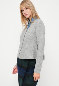 Sivka rebrasta jopica z dolgimi rokavi in odprtim sprednjim delom. Nosi se preko modre srajce s kratkimi rokavi. Usklajena z hlačami v karo vzorcu.