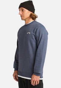 Sudadera azul de mezcla de algodón, con cuello redondo, puños acanalados y un logo bordado en el pecho. Llevada con un gorro negro.