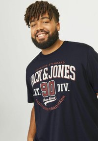 Mørkeblå kortærmet t-shirt lavet af bomuld, med iøjnefaldende hvide og bordeaux grafikker, hvor "Jack & Jones" og "90 Div." er trykt tydeligt.