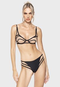 Vrouw met blond haar, gekleed in een zwarte doorschijnende beha met uitsnijdingen en bijpassende zwarte bikini-broekjes met meerdere zijkanten, tegen een grijze achtergrond.