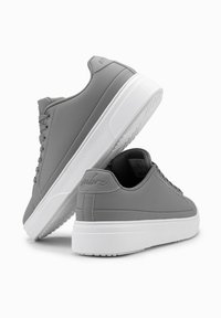 Des baskets grises en matériau lisse, avec une semelle en caoutchouc blanc, une forme arrondie et un design à lacets, comportant un détail de logo à l'arrière.