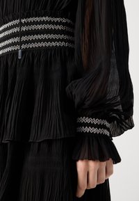 Sort plisseret kjole med gennemsigtige lange ærmer, hvid zigzagbroderet taljebånd og manchetter samt snor med metalspidser, der hænger ned foran.