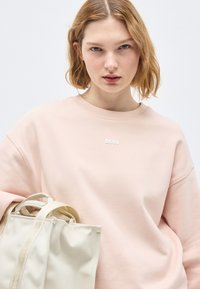 Νεαρή γυναίκα με κοντά ξανθά μαλλιά φοράει ανοιχτό ροζ φούτερ BOSS και κρατάει τσάντα tote σε κρεμ χρώμα με φόντο λευκό.