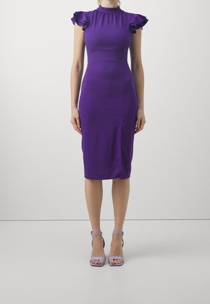 Nainen, joka pukeutuu polvipituiseen, istuvaan purppuranväriseen mekkoon, jossa on röyhelöiset hihojen liepeet, ja hänellä on jalassaan laventelinväriset nilkkaremmi-korkokengät. Hän seisoo yksinkertaisen vaalean taustan edessä.