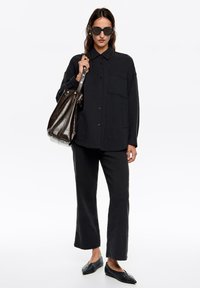 Camisa negra texturizada de botones con un bolsillo, combinada con pantalones negros de pierna ancha y zapatos planos negros. Los accesorios incluyen gafas de sol oscuras.