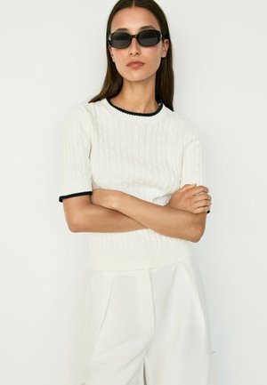 Frau mit langen dunklen Haaren, die schwarze Sonnenbrillen, ein weißes kurzärmliges Stricktop mit dunklem Besatz und weiße Hosen trägt, steht mit verschränkten Armen.