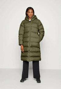 Naine seisab, seljas pikk oliiviroheline sulejope kapuutsiga, mustad püksid ja mustad tossud, neutraalse tausta ees.