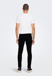 Camiseta blanca de manga corta, jeans ajustados negros y zapatillas negras con detalles morados, vistas desde atrás sobre un fondo claro.