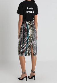 T-shirt noir avec le texte "RIRE PLUS FORT" au dos, associé à une jupe midi multicolore en sequins avec des rayures verticales et une fente sur la cuisse.
