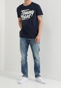 Mörkblå bomullströja med vit "Tommy Jeans"-logotyp, kombinerad med ljusblå denimjeans och vita sneakers. Enkelt, avslappnat design.