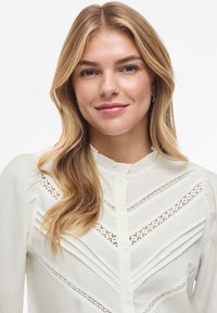 Balta palaidinė su raukšlėtu apykakliu, dekoratyvine nėrinių detalė ir sagomis priekyje. Turi ilgomis rankovėmis ir minkštą, tekstūruotą audinį.