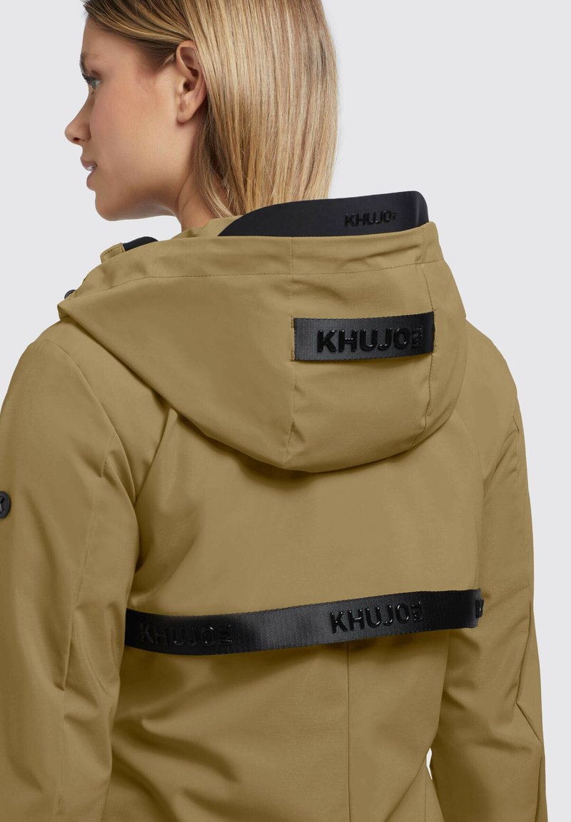 Veste khaki à capuche avec une finition mate, présentant une sangle de logo noire dans le dos et un col haut pour une protection supplémentaire.