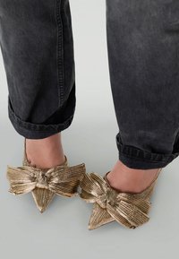 Chaussures à talons hauts dorées avec un grand nœud, bout pointu et finition texturée, portées avec un jean noir retroussé.