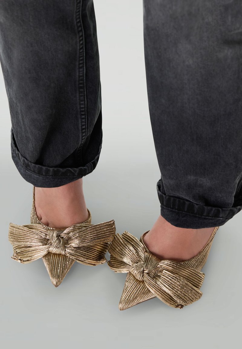 Chaussures à talons hauts dorées avec un grand nœud, bout pointu et finition texturée, portées avec un jean noir retroussé.