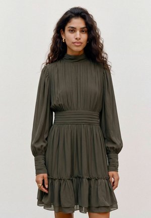 Un mannequin aux longs cheveux foncés se tient dans un studio, vêtu d’une robe plissée vert olive foncé à manches bouffantes, avec un col montant et un ourlet à volants.