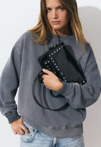 Une femme portant un sweat gris patiné et un jean clair tient un sac à main en cuir noir clouté près de sa poitrine.