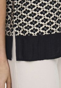 Blouse à motifs noir et blanc avec un design floral et une accentuation noire horizontale à l'ourlet ; le tissu semble léger et texturé.
