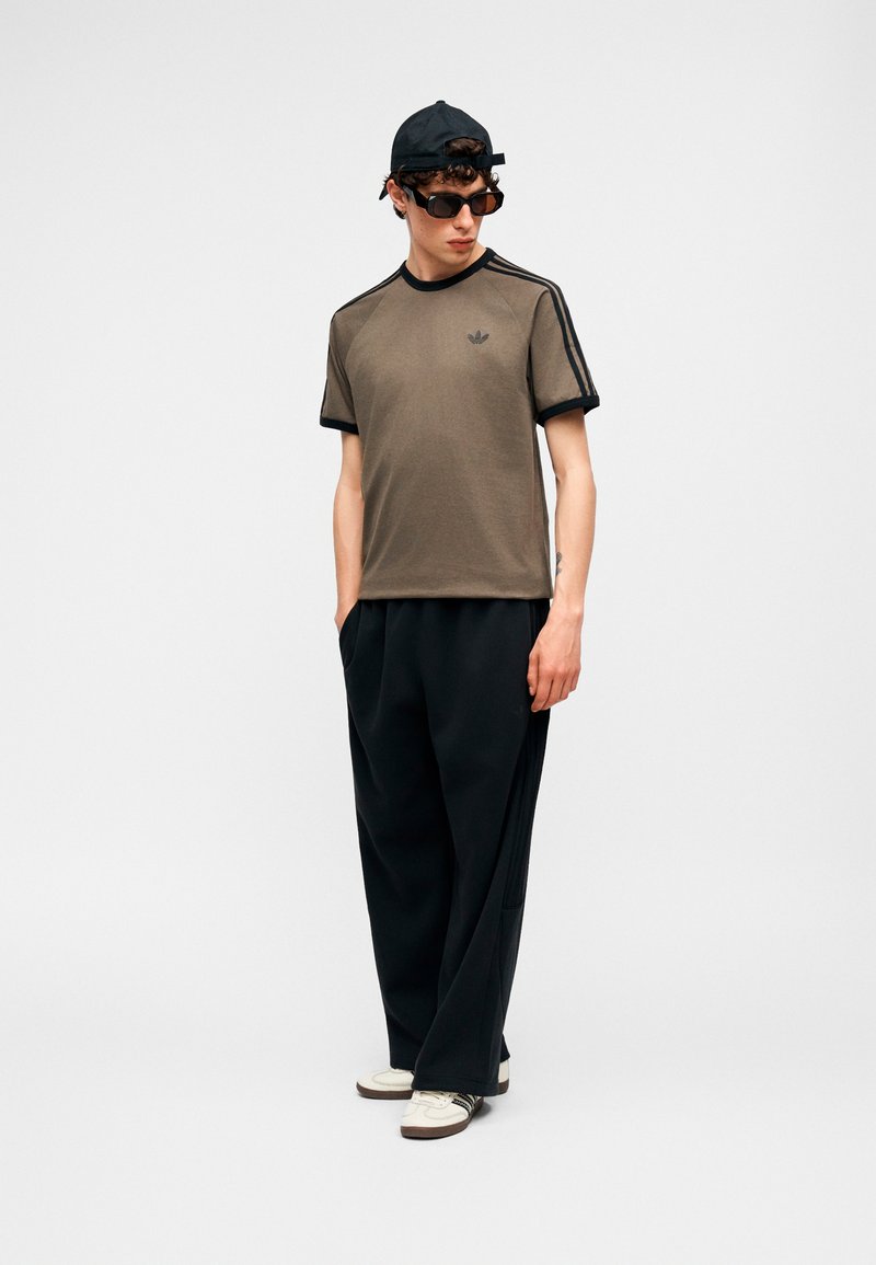 Ung mand iført solbriller, sort kasket, brun Adidas t-shirt, sorte brede bukser, hvide sneakers, stående med hænderne i lommerne, kiggende ned mod venstre.