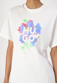 Tricou alb cu mânecă scurtă, având un design abstract colorat în nuanțe de albastru, roșu și verde, cu textul "HUGO" scris cu litere albe îngroșate.