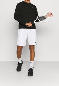 Černá mikina s kulatým výstřihem, bílé šortky s kapsami, černé tenisky a bílé sportovní ponožky. Drží bílou tenisovou raketu.