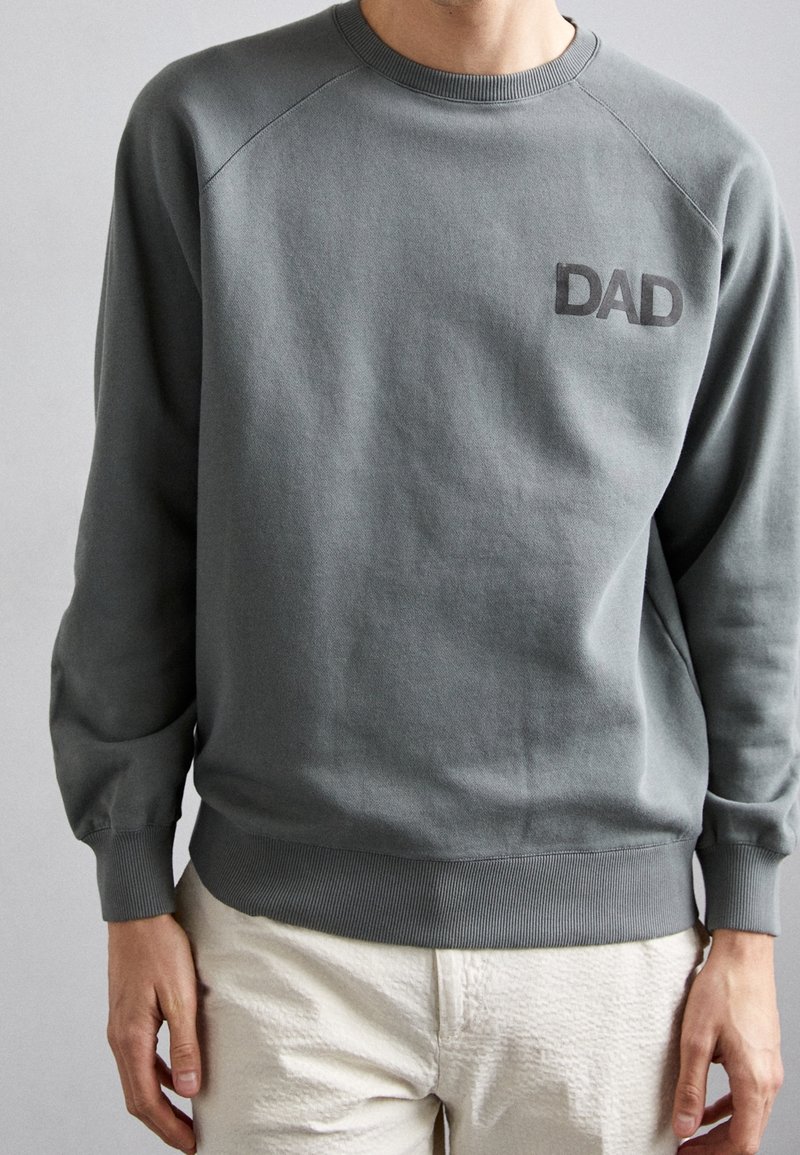 Muž v šedé mikině s nápisem "DAD" na hrudi a krémových kalhotách, stojící na jednobarevném pozadí.
